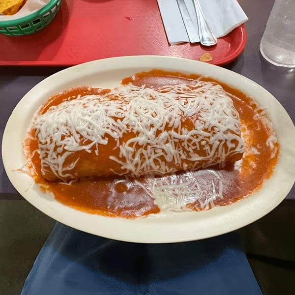 A large, saucy burrito covered in melted white cheese on a white oval plate.